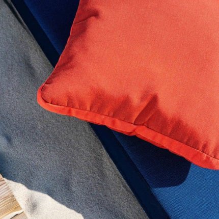 zbliżenie na trzy warstwy tkanin Sunbrella o różnych kolorach: żywą poduszkę w kolorze koralowym, leżącą na intensywnie niebieskim materiale, obok ciemnoszarej, teksturowanej tkaniny