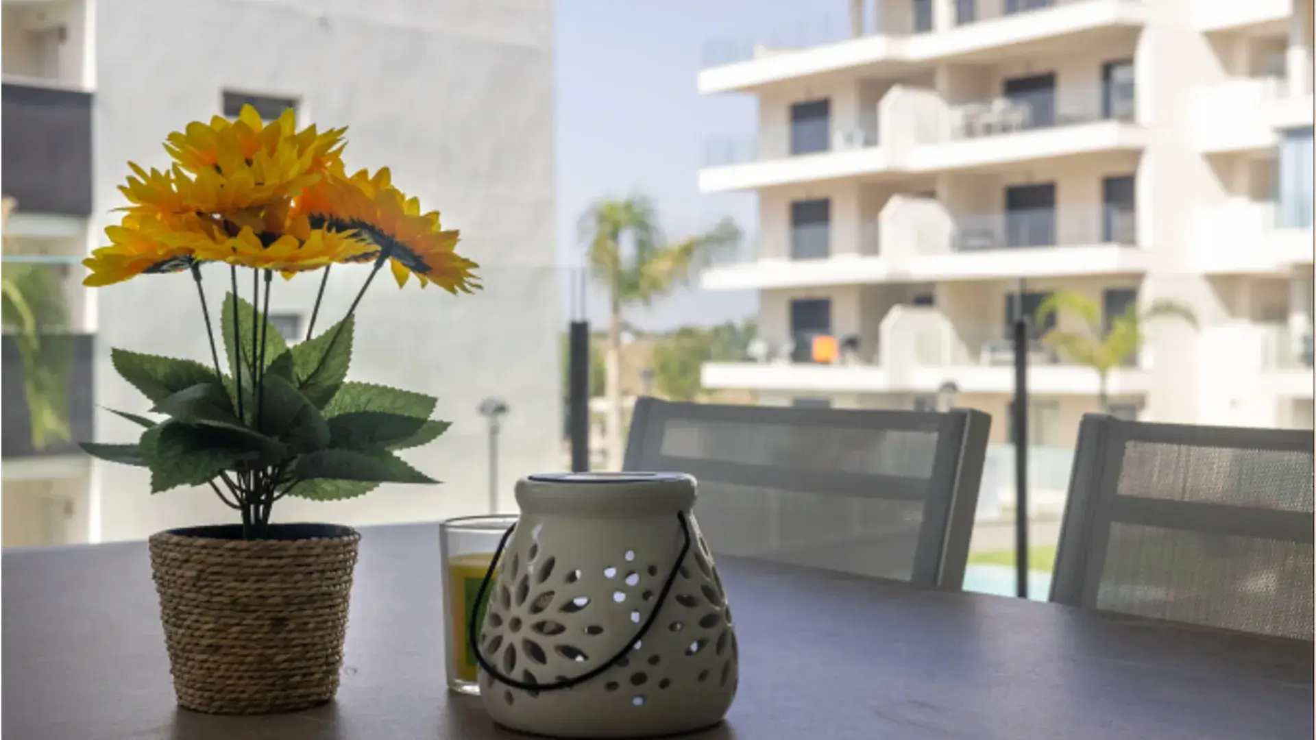 Jak urządzić balkon w bloku - Stół balkonowy z dekoracją – sztuczne słoneczniki i świecznik na tle nowoczesnych budynków