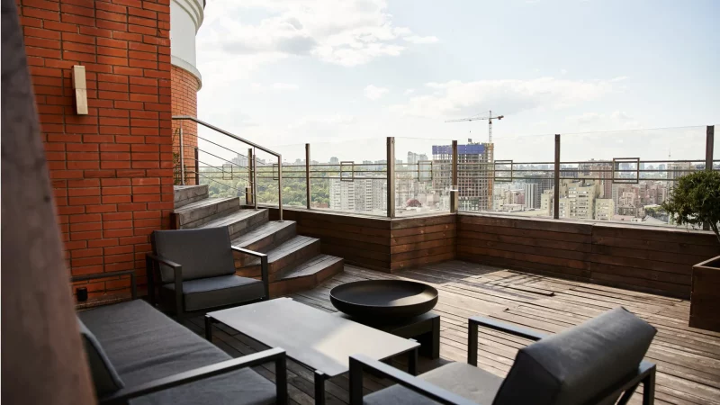 Jak ładnie i nowocześnie urządzić balkon? Narożny balkon z widokiem na miasto i zestawem wypoczynkowym z czarnymi fotelami