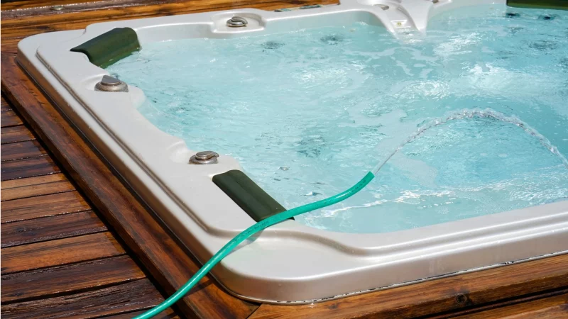Jak dbać o wodę w jacuzzi ogrodowym? Ogrodowe jacuzzi obudowane drewnem, z dyszami hydromasażu i zielonym wężem ogrodowym, widać krystalicznie czystą wodę oraz delikatnie unoszącą się pianę