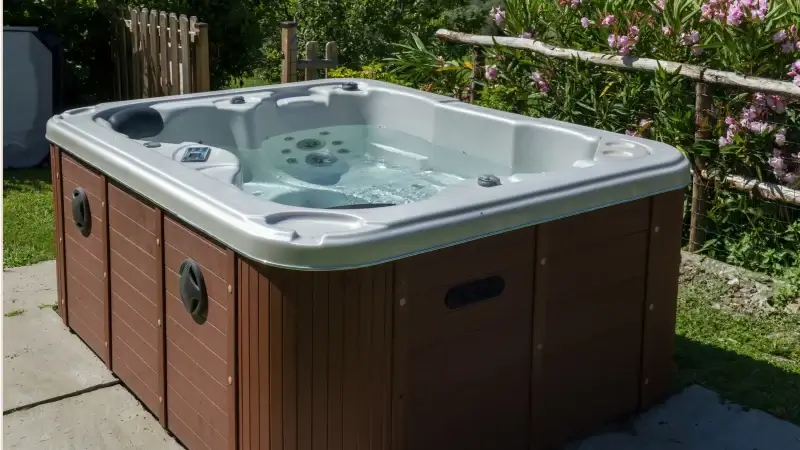 Nowoczesne jacuzzi ogrodowe z drewnianą obudową i srebrną niecką, ustawione na kamiennej posadzce w otoczeniu zieleni i kwitnących krzewów.