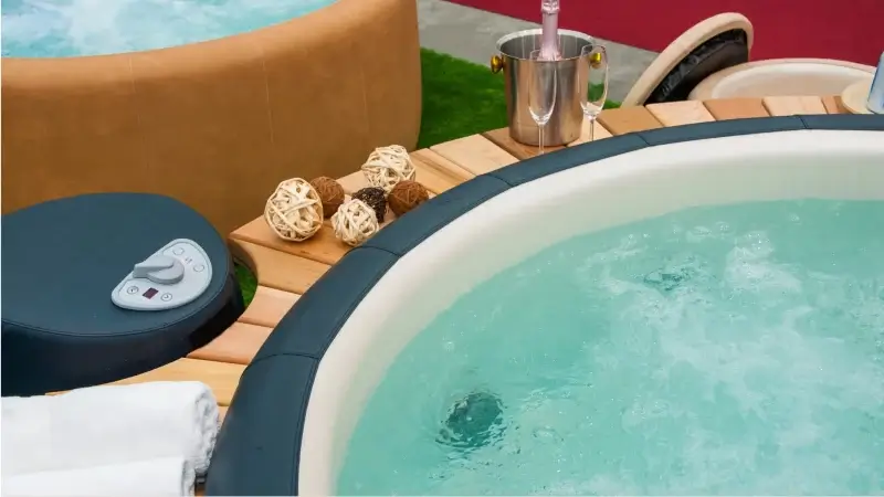 Zbliżenie na okrągłe jacuzzi z podgrzewaną wodą, obok leżą ręczniki, dekoracyjne kule i stoją kieliszki do szampana z butelką w chłodziarce