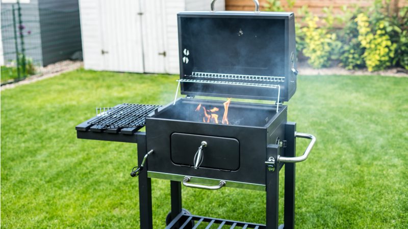 Jak urządzić taras w stylu hampton? Czarny grill ogrodowy z otwartą pokrywą i płomieniem wewnątrz, ustawiony na trawniku w ogrodzie