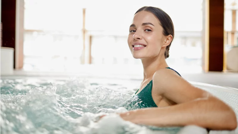 Czy jacuzzi jest zdrowe? Jak długo można w nim przebywać? Uśmiechnięta kobieta w kostiumie kąpielowym korzystająca z jacuzzi z bąbelkami, wnętrze spa z jasnym światłem w tle.