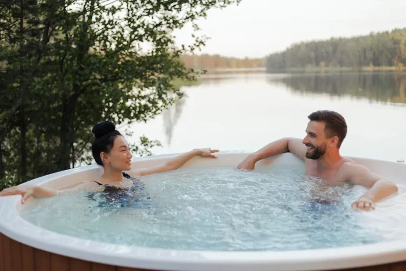 Czy jacuzzi jest zdrowe? Jak długo można w nim przebywać? Para relaksująca się w jacuzzi ustawionym nad jeziorem, z widokiem na wodę i drzewa, atmosfera wypoczynku na świeżym powietrzu.