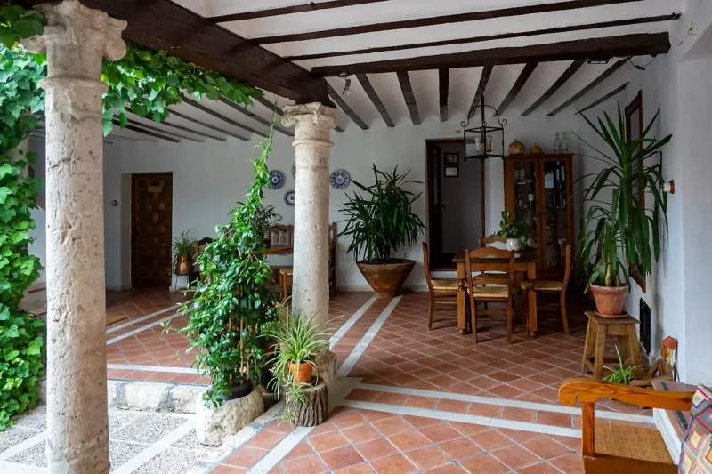 Jak urządzić taras i ogród w stylu hiszpańskim? Hiszpańskie patio z kamiennymi kolumnami, bielonymi ścianami i drewnianymi belkami stropowymi, urządzone roślinami w donicach i rustykalnymi meblami z naturalnego drewna.